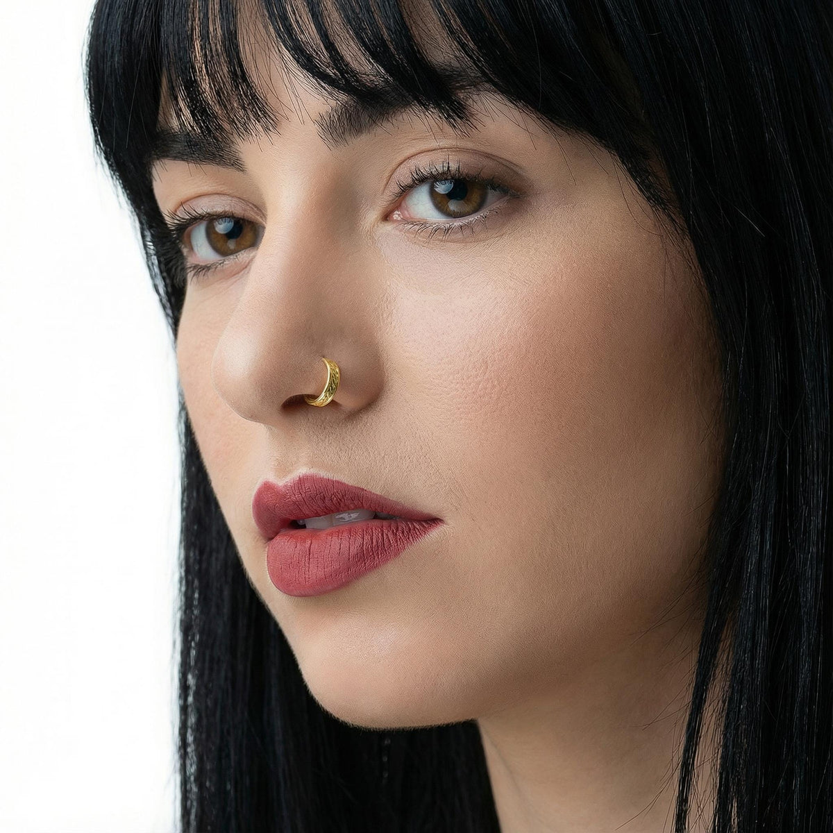 A close-up portrait of a woman with dark hair and a nose ring, looking directly at the camera with a serious expression.