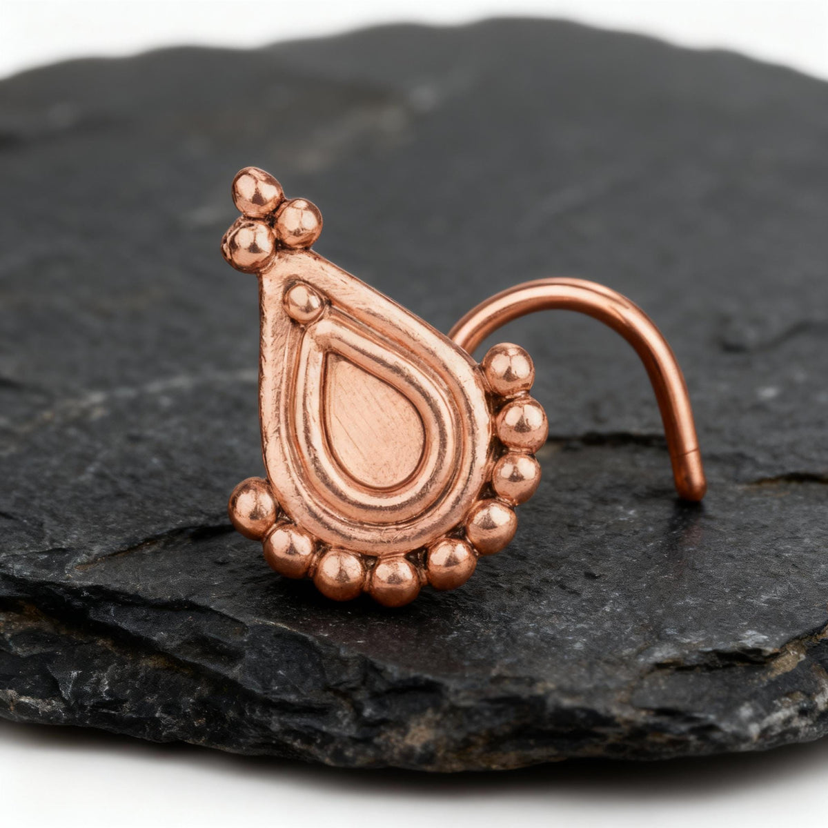 Rose Gold Indian Design Nose Stud: A copper-colored nose ring with a decorative pendant is displayed on a dark surface.