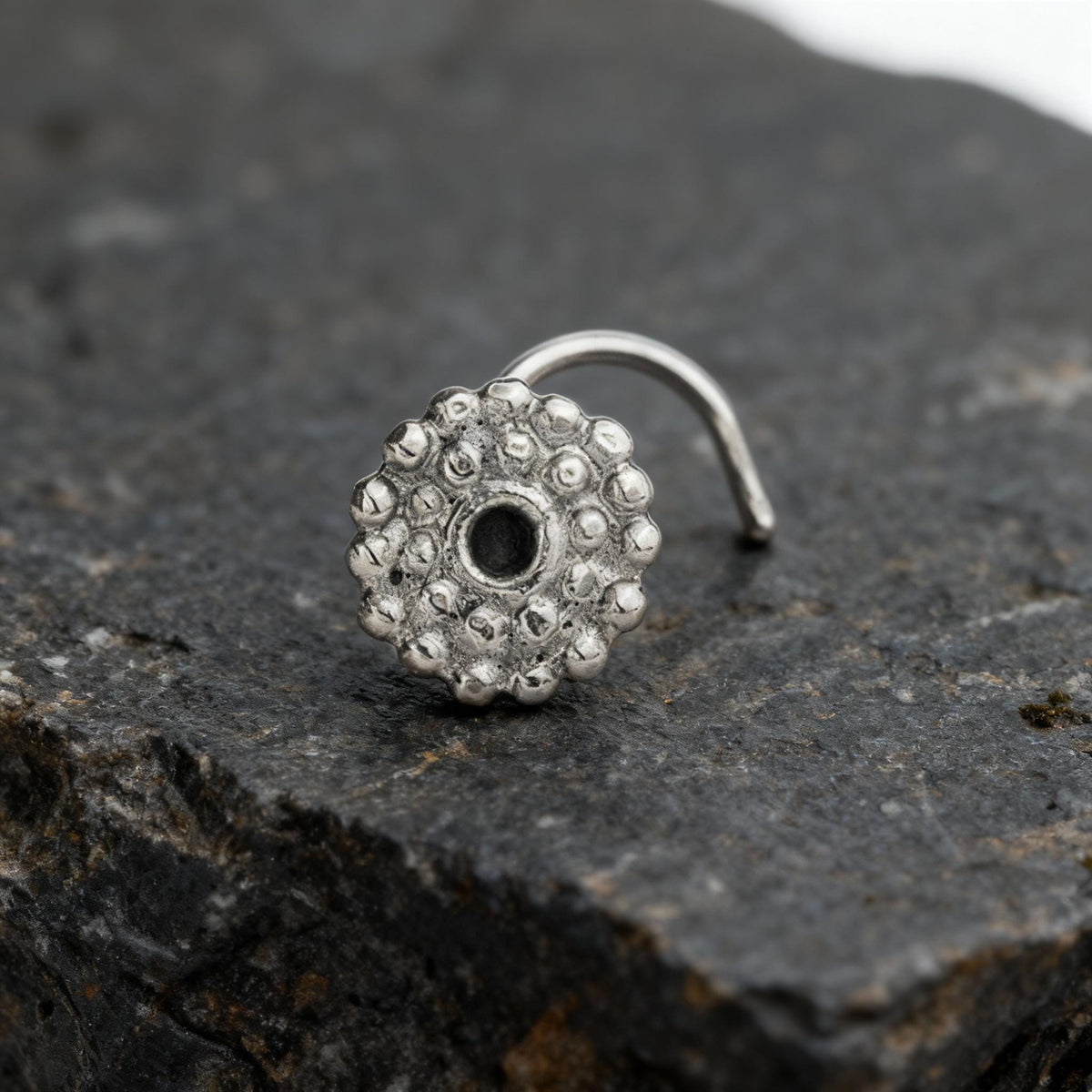 Indian Nose Stud: A silver-colored nose ring with a circular design sits on a dark surface.