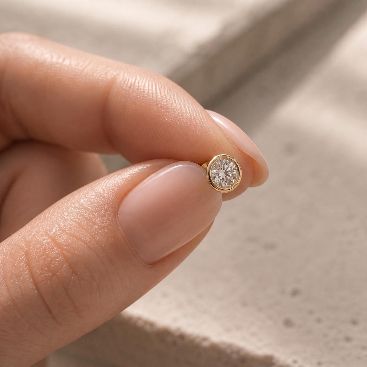 A person's hand holding a small gold-colored diamond earring with a round brilliant-cut diamond.