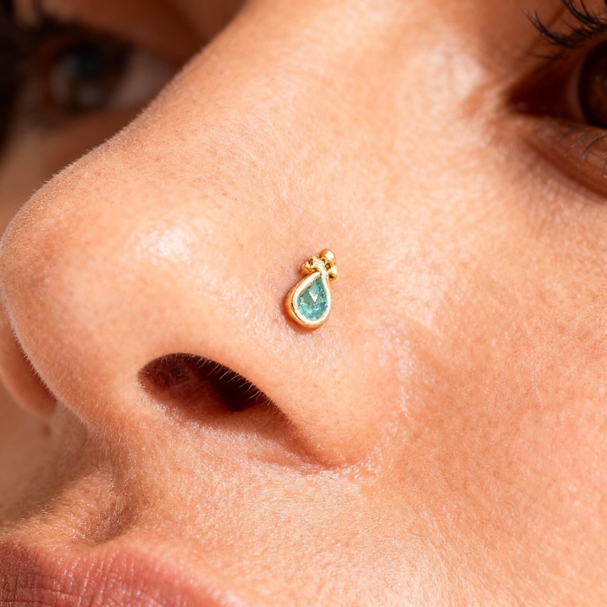 A close-up of a person's nose with a small gold-colored earring featuring a teardrop-shaped gemstone protruding from it.