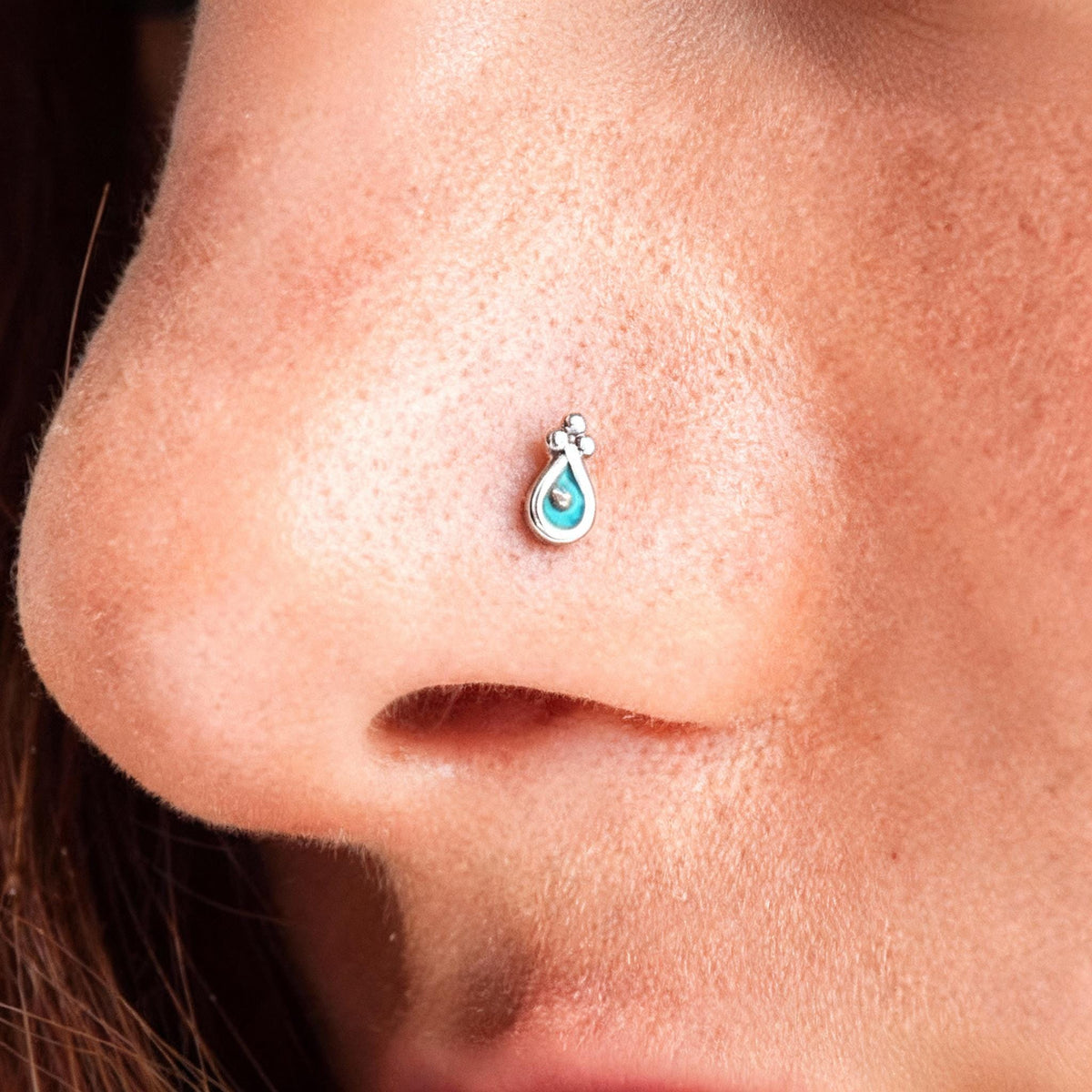 A close-up of a person's nose with a small, turquoise-colored piercing on the nostril.