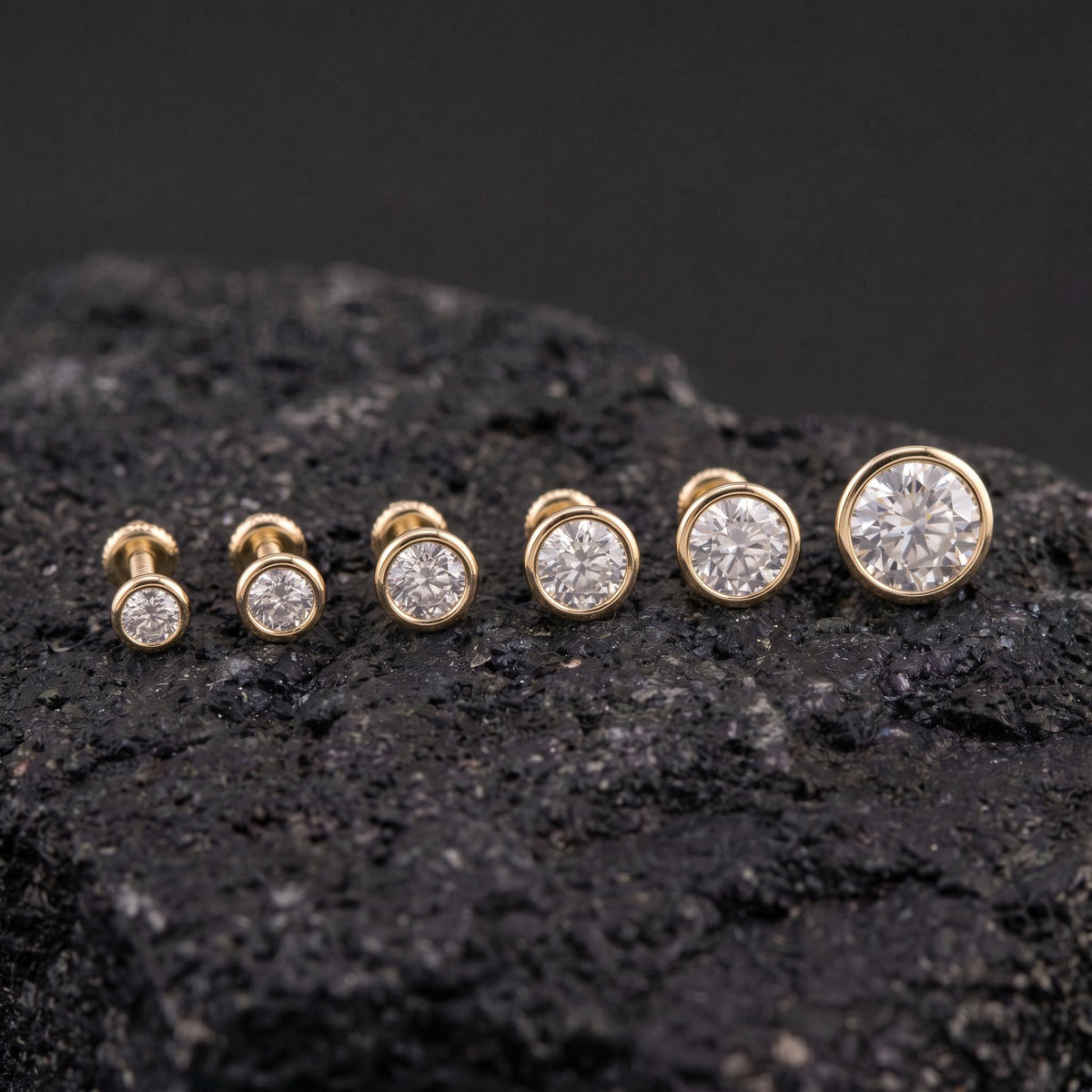 A collection of six gold-colored diamond stud earrings with large, clear gemstones is displayed on a dark, rocky surface.