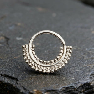 A silver-colored metal piercing ring with a decorative design on a dark surface.