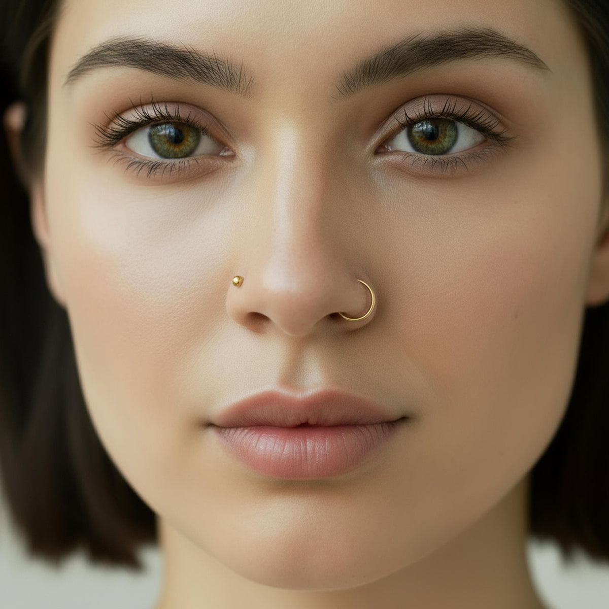 A close-up portrait of a woman with dark hair and a nose piercing, looking directly at the camera with a neutral expression.