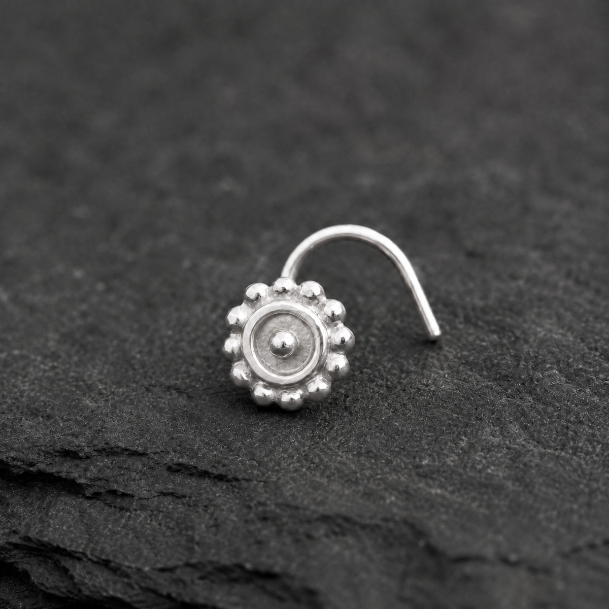 A silver nose stud with a round center and small beads is resting on a dark surface.