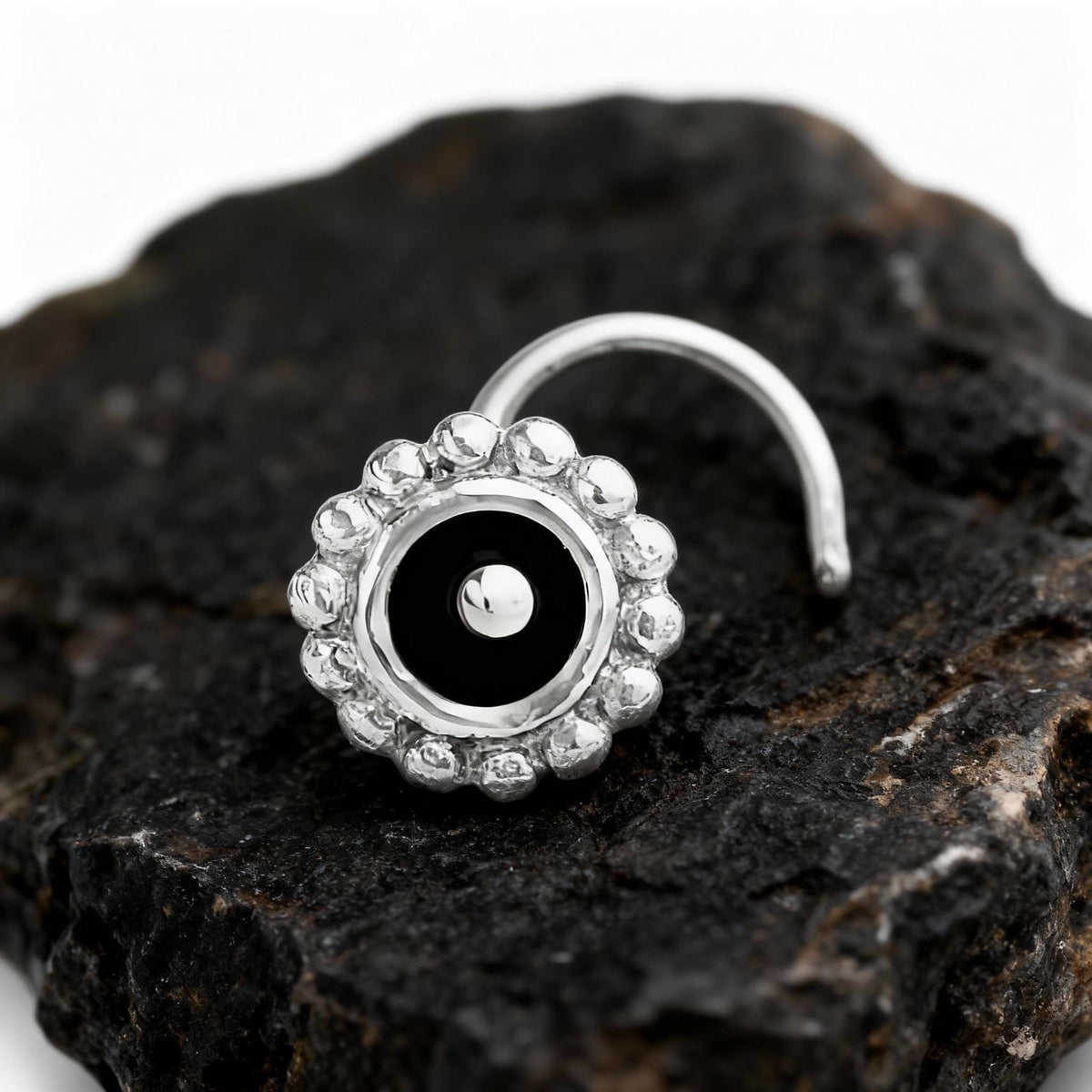 A silver nose ring with a black center and small white stones is resting on a dark, rocky surface.