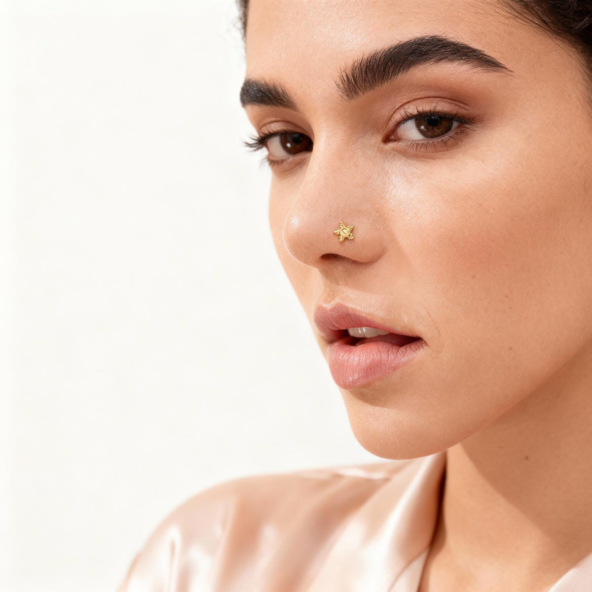A close-up portrait of a woman with a gold star-shaped nose stud, wearing a pink satin blouse.
