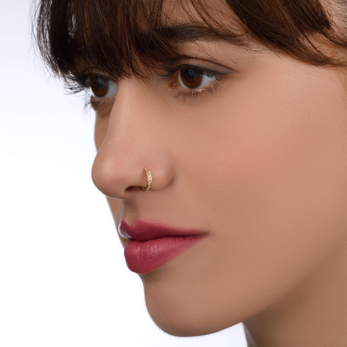 A close-up portrait of a woman with dark hair and a nose ring, looking off to the side with a neutral expression.