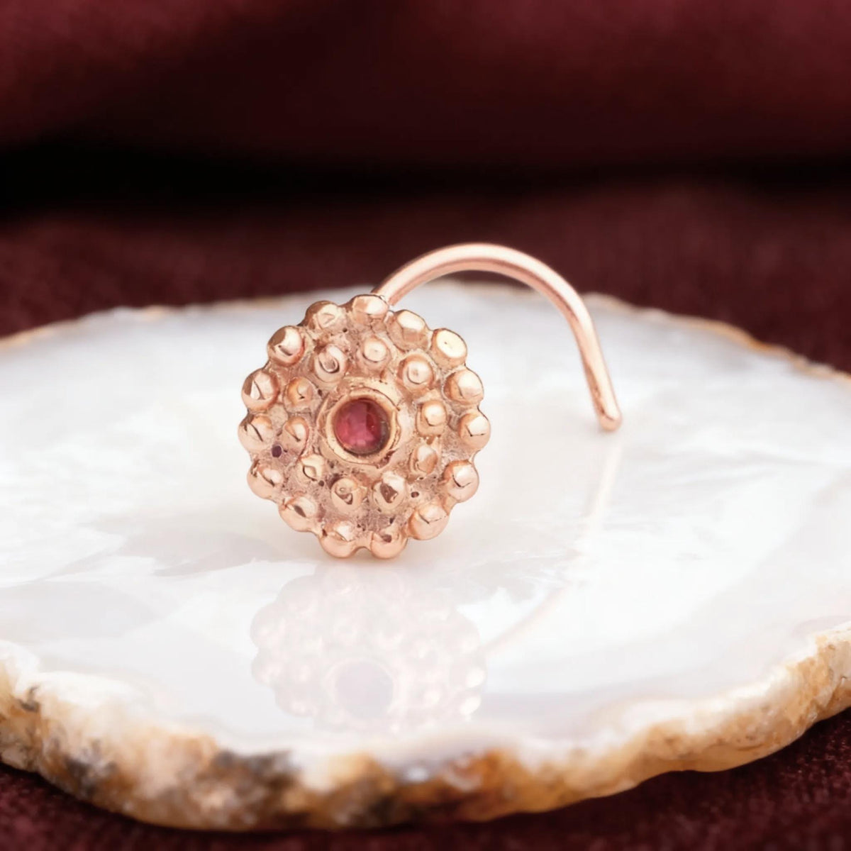 A gold-colored nose ring with a red gemstone is displayed on a white surface against a burgundy background.