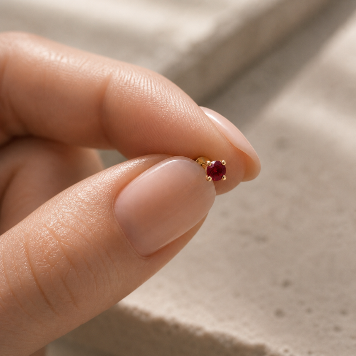 A person's hand holding a small gold-colored earring with a red gemstone.