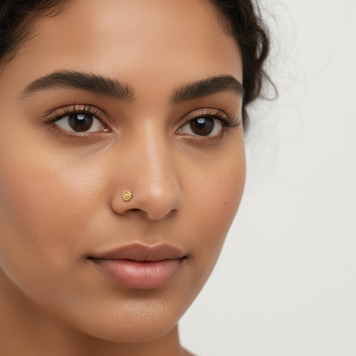 A close-up portrait of a woman with dark hair and a gold nose stud.