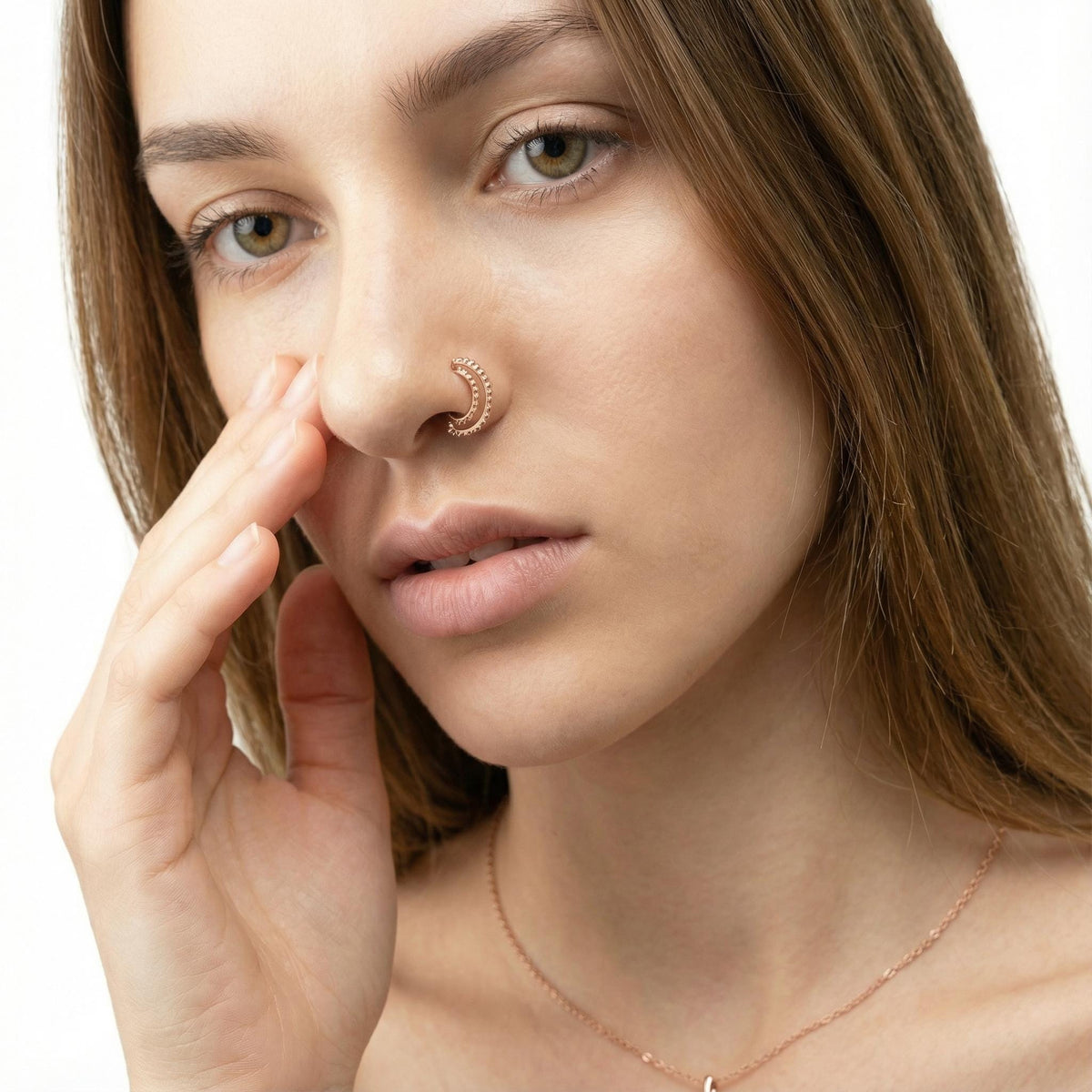 A close-up portrait of a woman with long brown hair, wearing a nose ring and a necklace. She is touching her nose with her finger, and her gaze is directed off to the side.