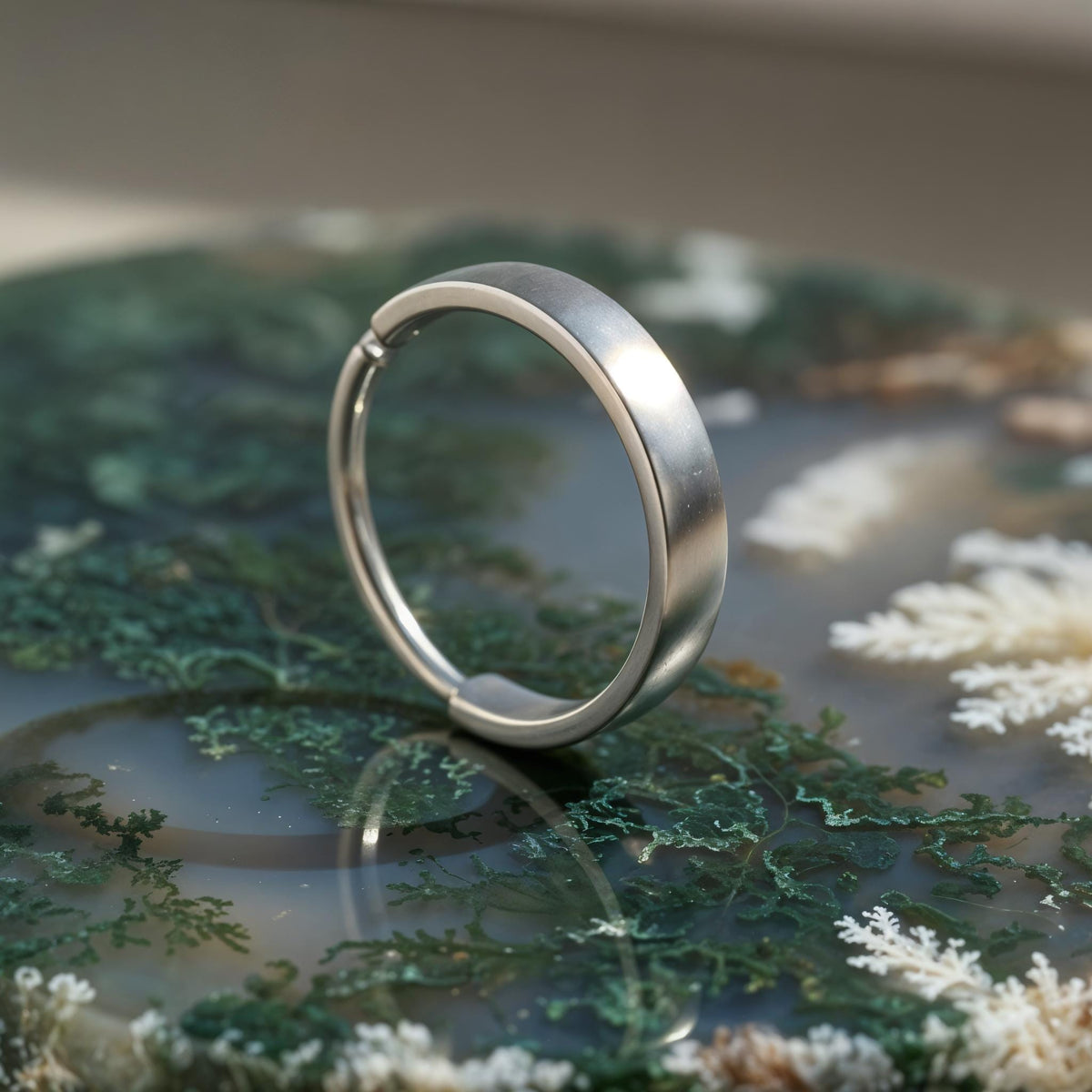 A silver ring with a curved band is resting on a surface covered in green moss and white flowers.