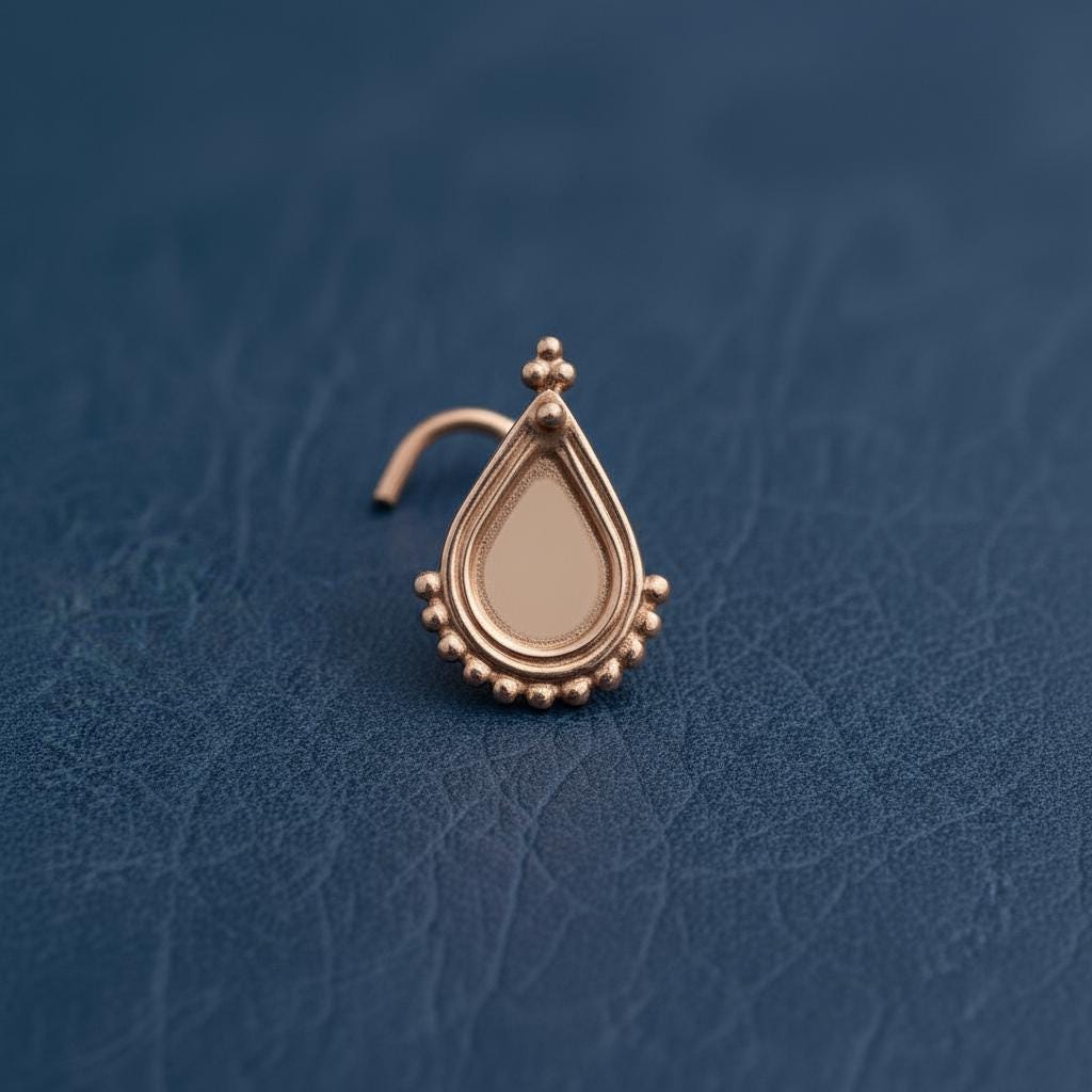 A gold-colored nose stud with a teardrop-shaped design and small beads around the base is displayed against a blue background.