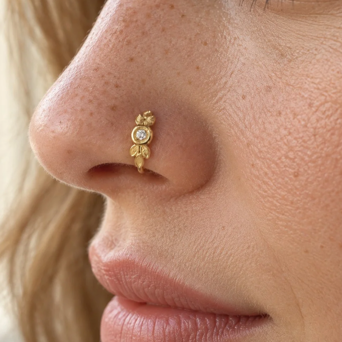 A close-up of a person's nose with a gold-colored nose ring featuring a small diamond and leaf-shaped designs.