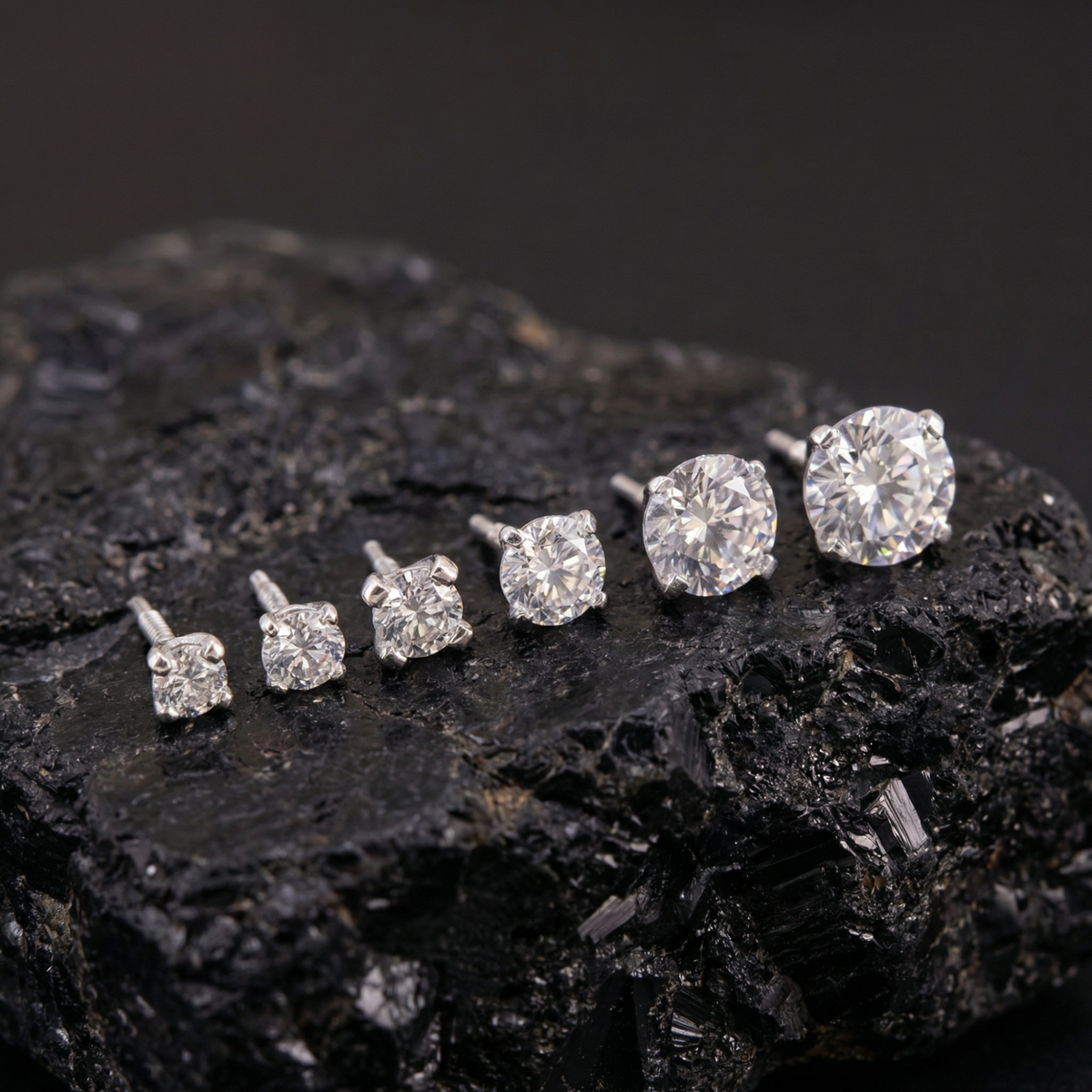 A collection of six diamond earrings in various sizes, ranging from small to large, displayed on a black rock.