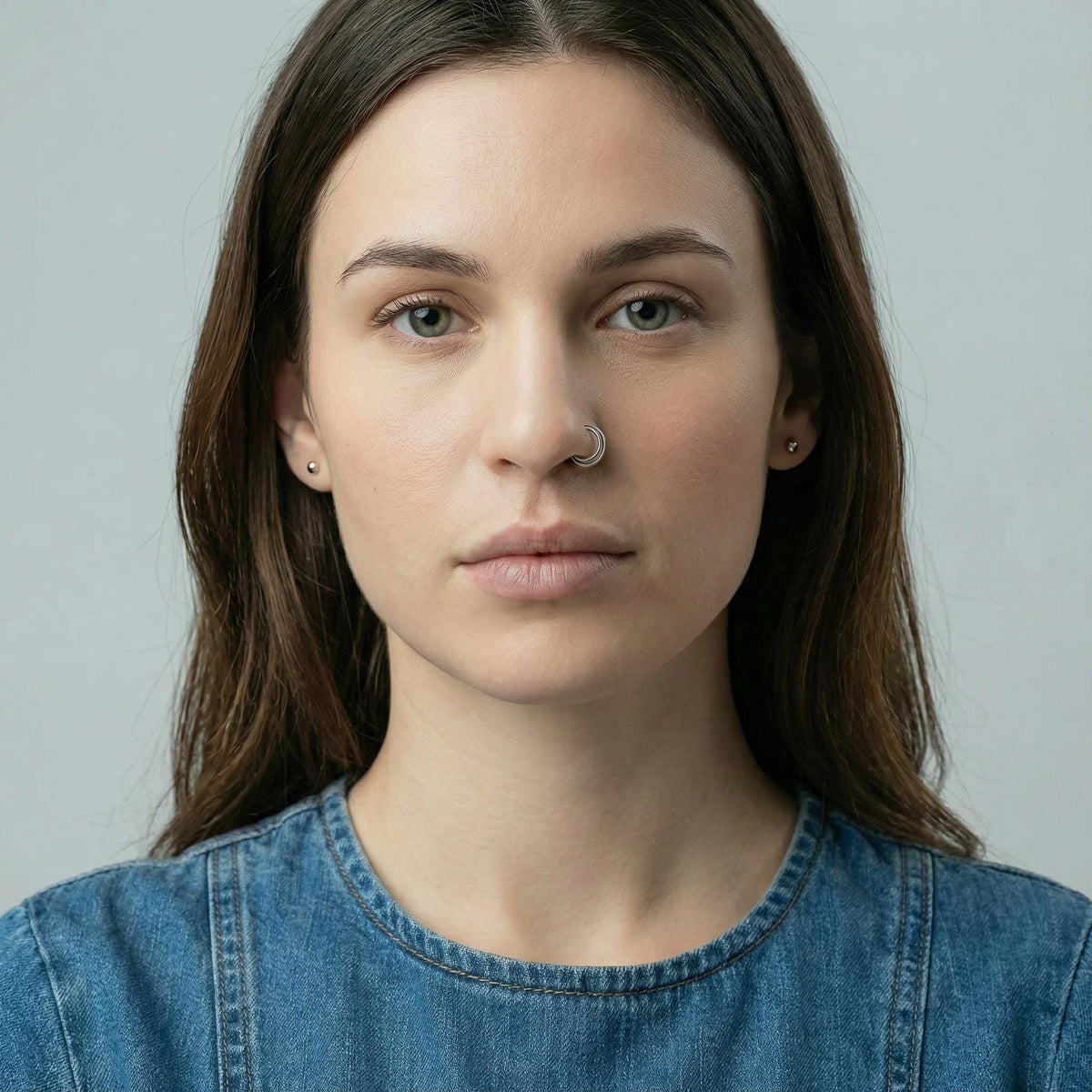 A portrait of a young woman with long brown hair, wearing a denim shirt and a nose hoop ring.