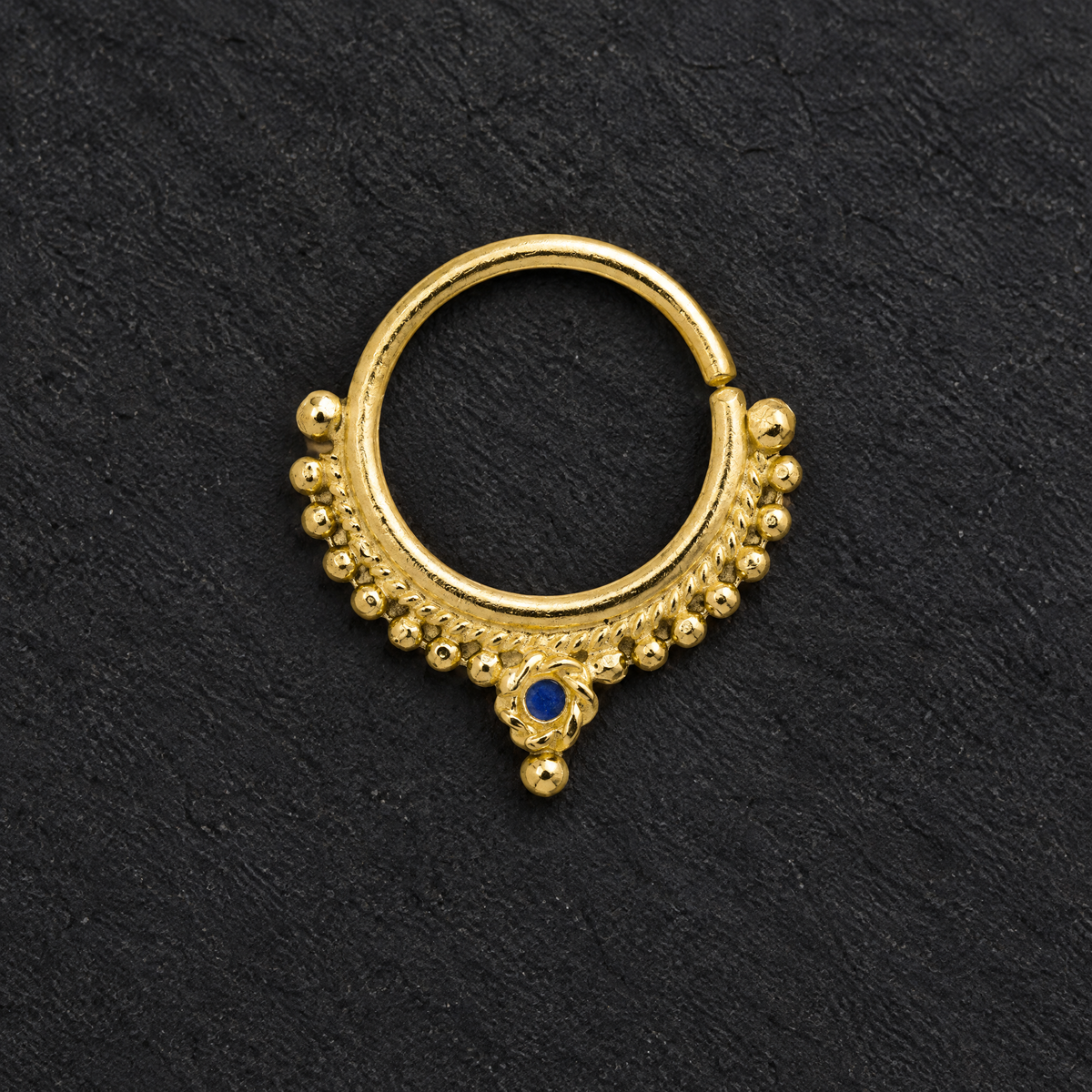A gold-colored nose ring with a blue gemstone is shown against a dark background.