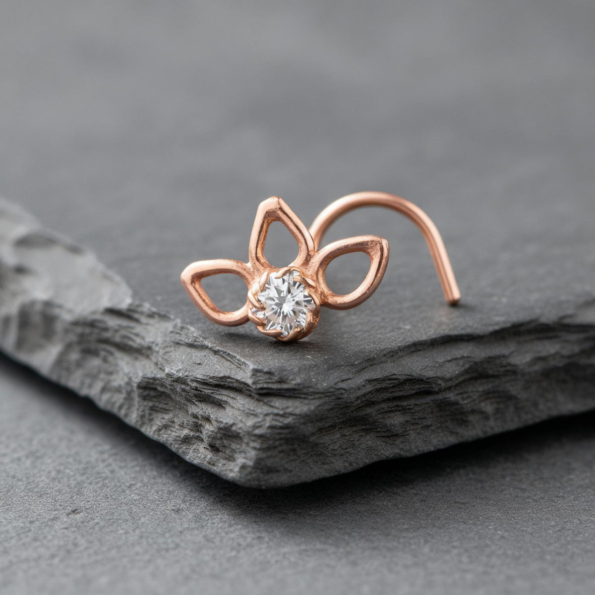 A rose gold nose ring with a clear gemstone is displayed on a gray rock.