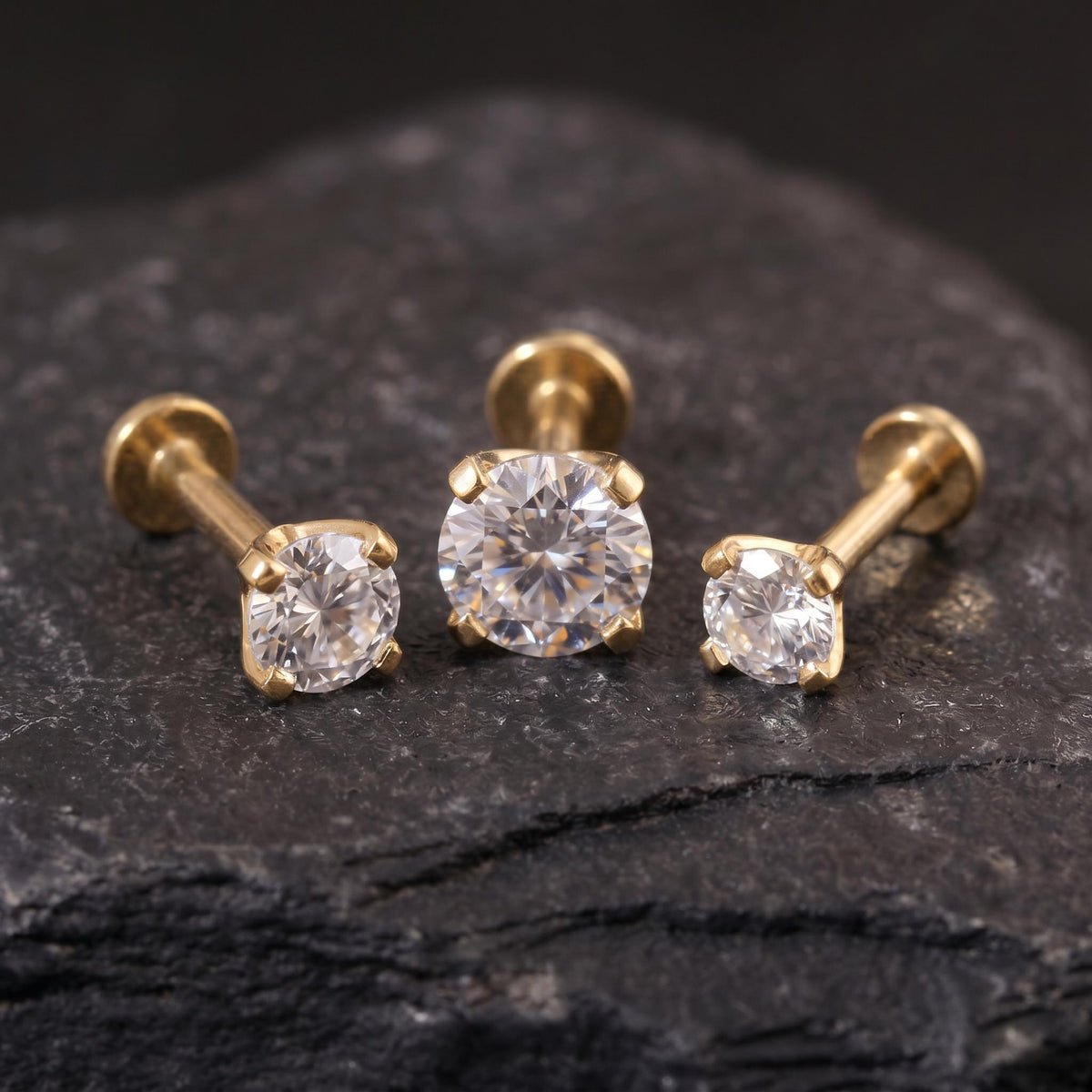 Three gold-colored diamond earrings are displayed on a dark surface, with the largest earring in the center and the other two smaller ones on either side.