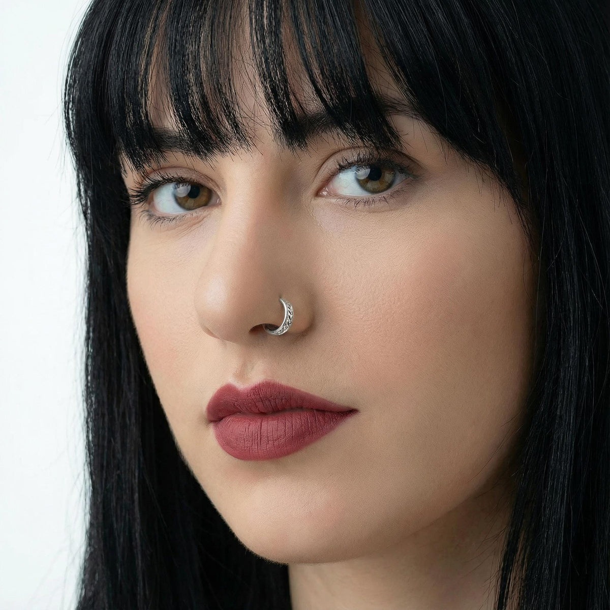 A close-up portrait of a woman with dark hair and a nose ring, looking directly at the camera with a serious expression.