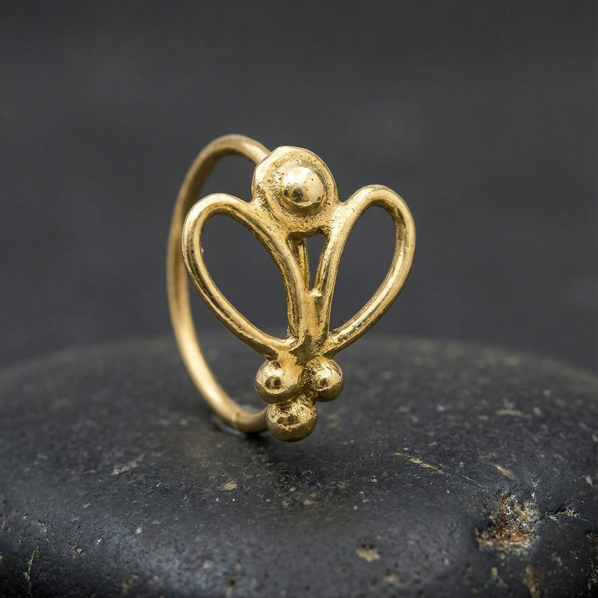 A gold-colored metal nose ring with an intricate floral design is displayed on a black surface.