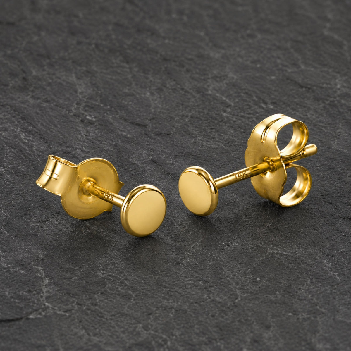Two gold-colored stud earrings with round backings are displayed on a dark surface.