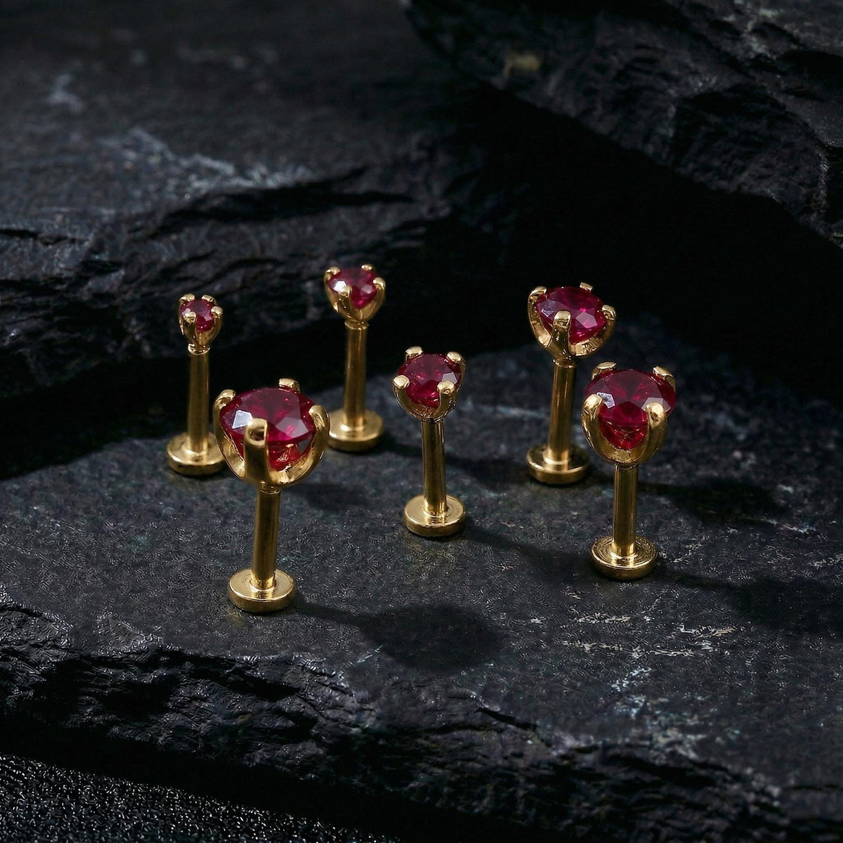 A group of six gold-colored earring posts with red gemstones, arranged on a dark surface.