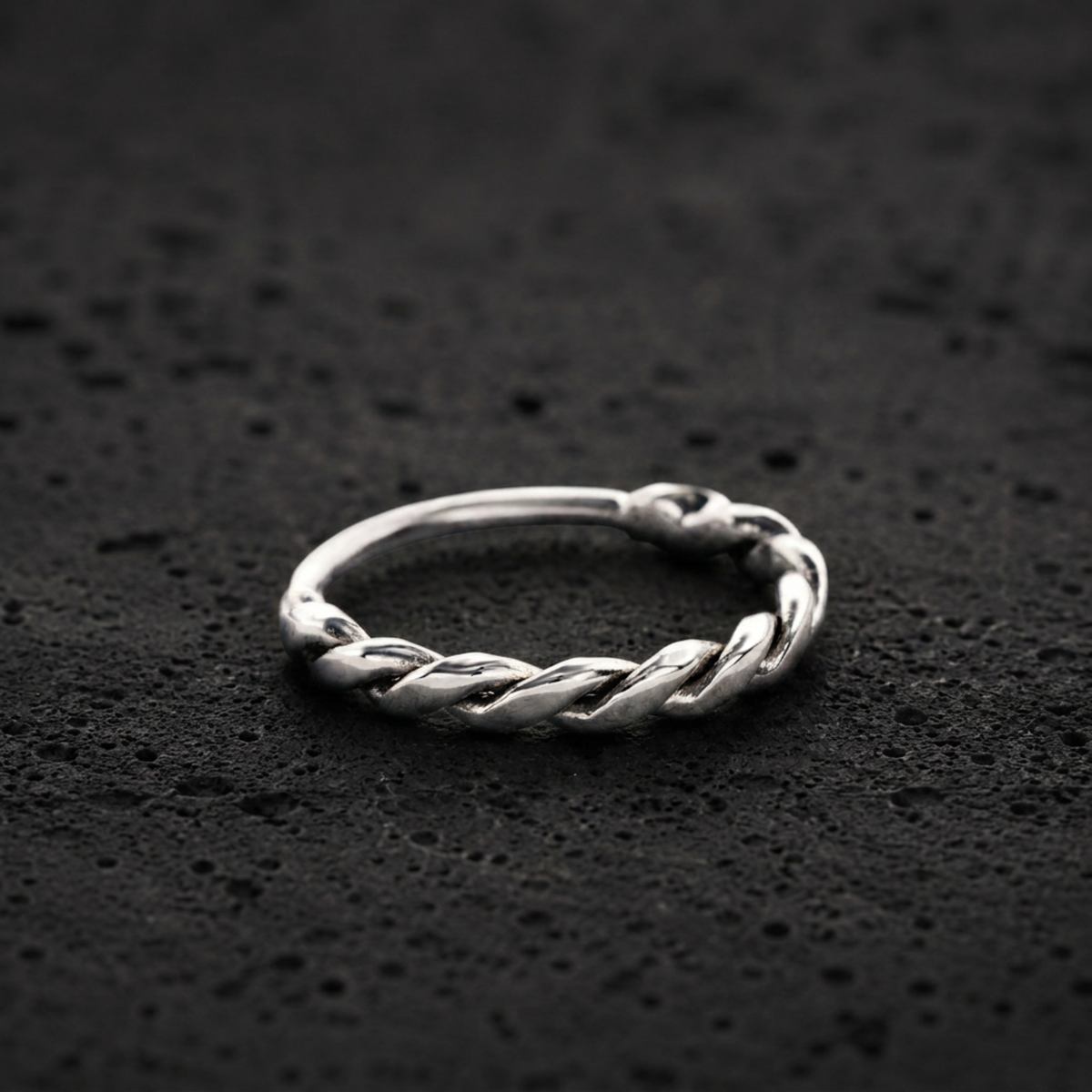 A silver ring with a twisted design sits on a dark, textured surface.