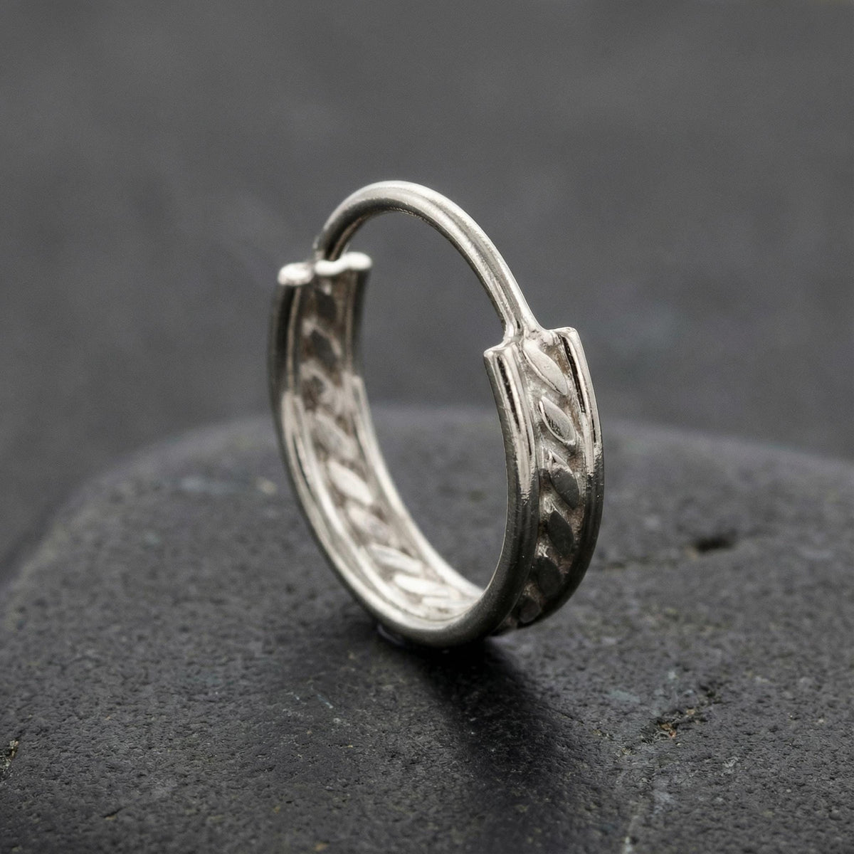 A silver hoop earring with a decorative design rests on a dark surface.