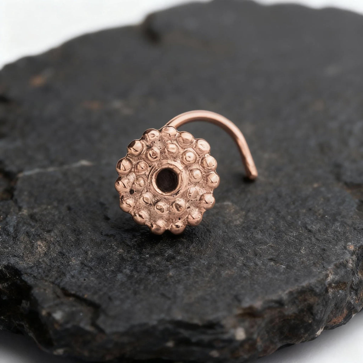 A copper-colored metal nose ring with a round hole is resting on a dark, textured surface.