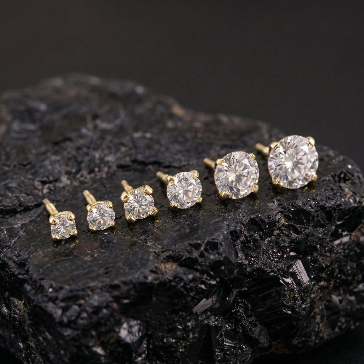 A collection of six gold earrings with large, clear diamonds set in them, arranged on a black rock.