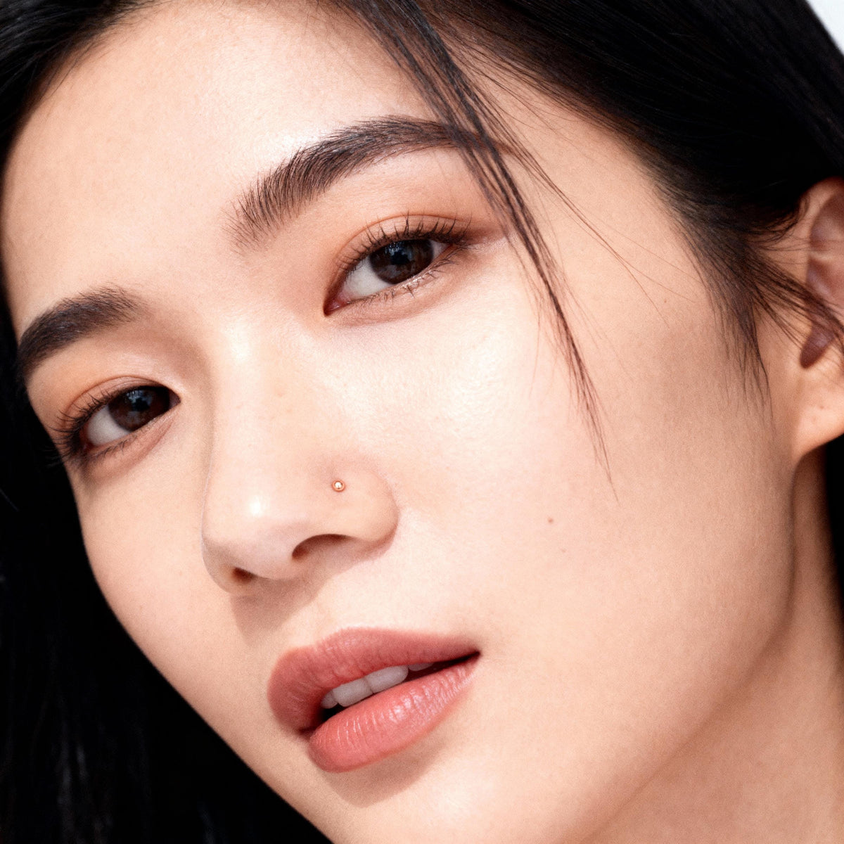 A close-up portrait of a young Asian woman with dark hair and a nose piercing, looking directly at the camera with a neutral expression.