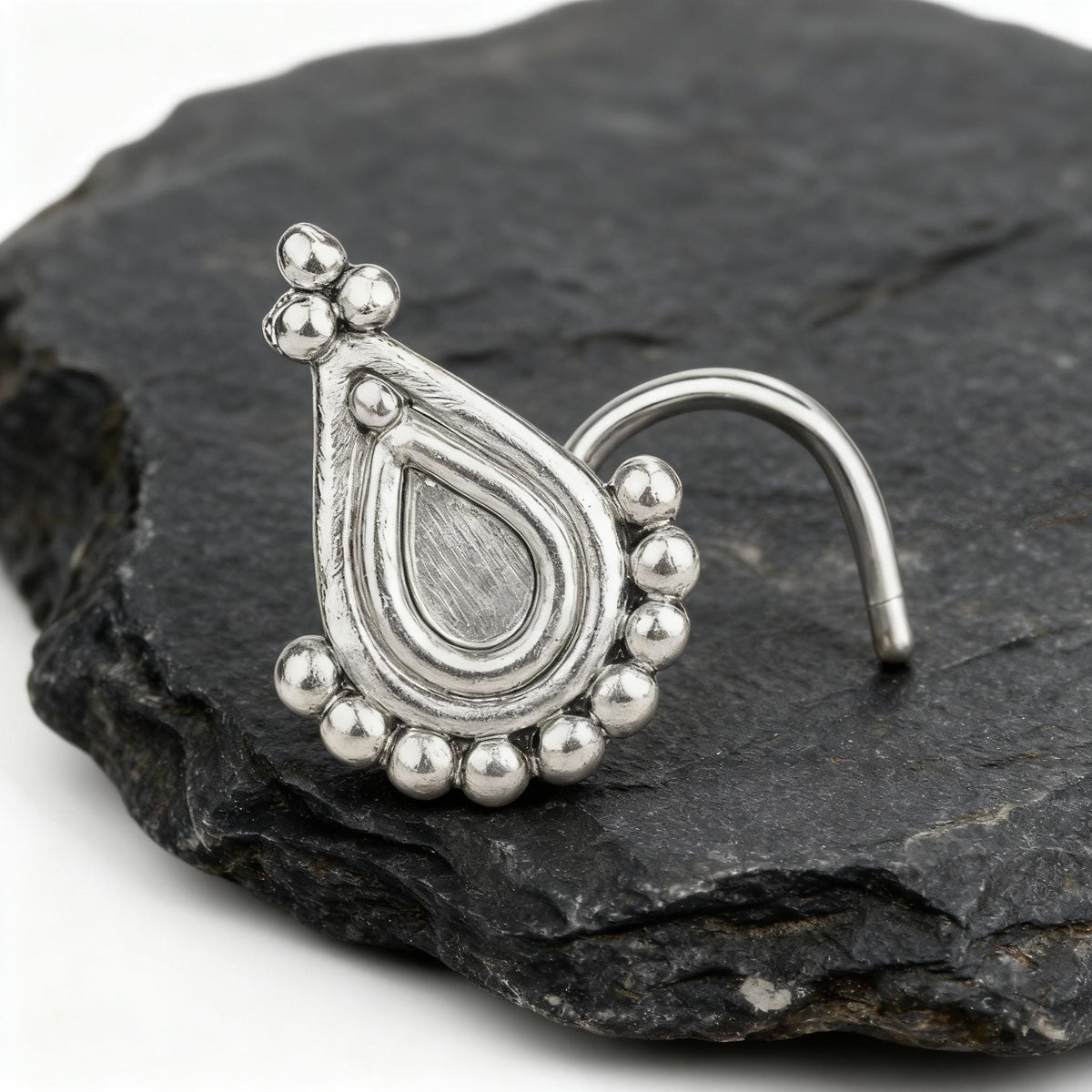 A silver nose ring with a decorative pendant is displayed on a dark gray rock.