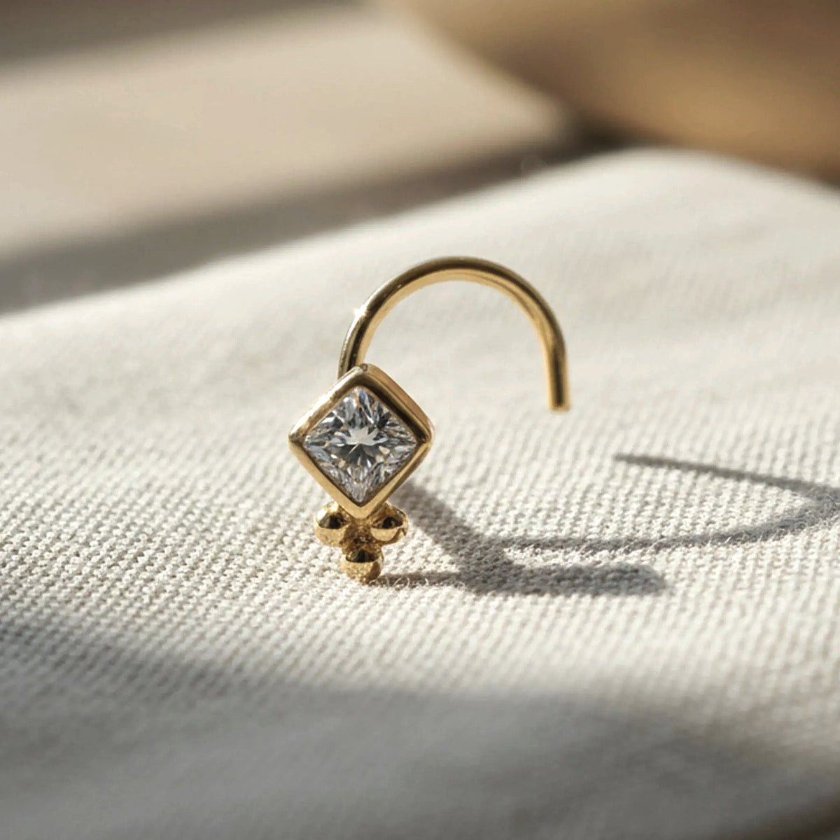 Diamond Nose Stud: A gold nose stud with a diamond-like center stone is displayed on a white fabric surface.