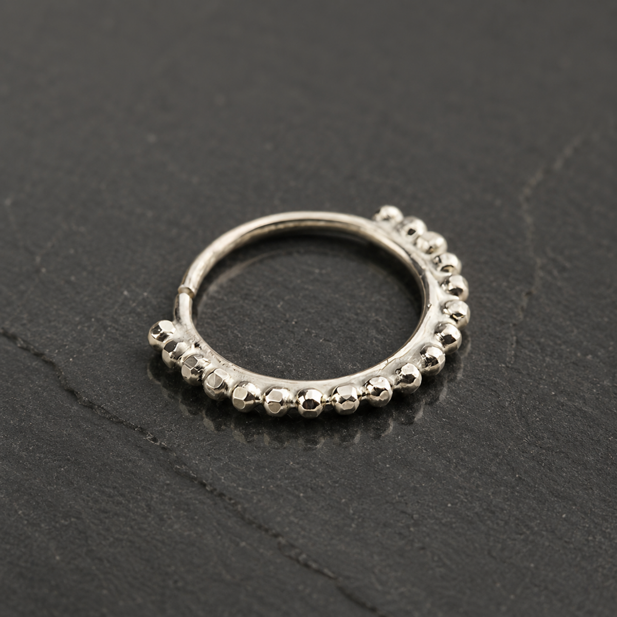 A silver-colored metal ring with a row of small, round beads or stones along the edge, resting on a dark surface.
