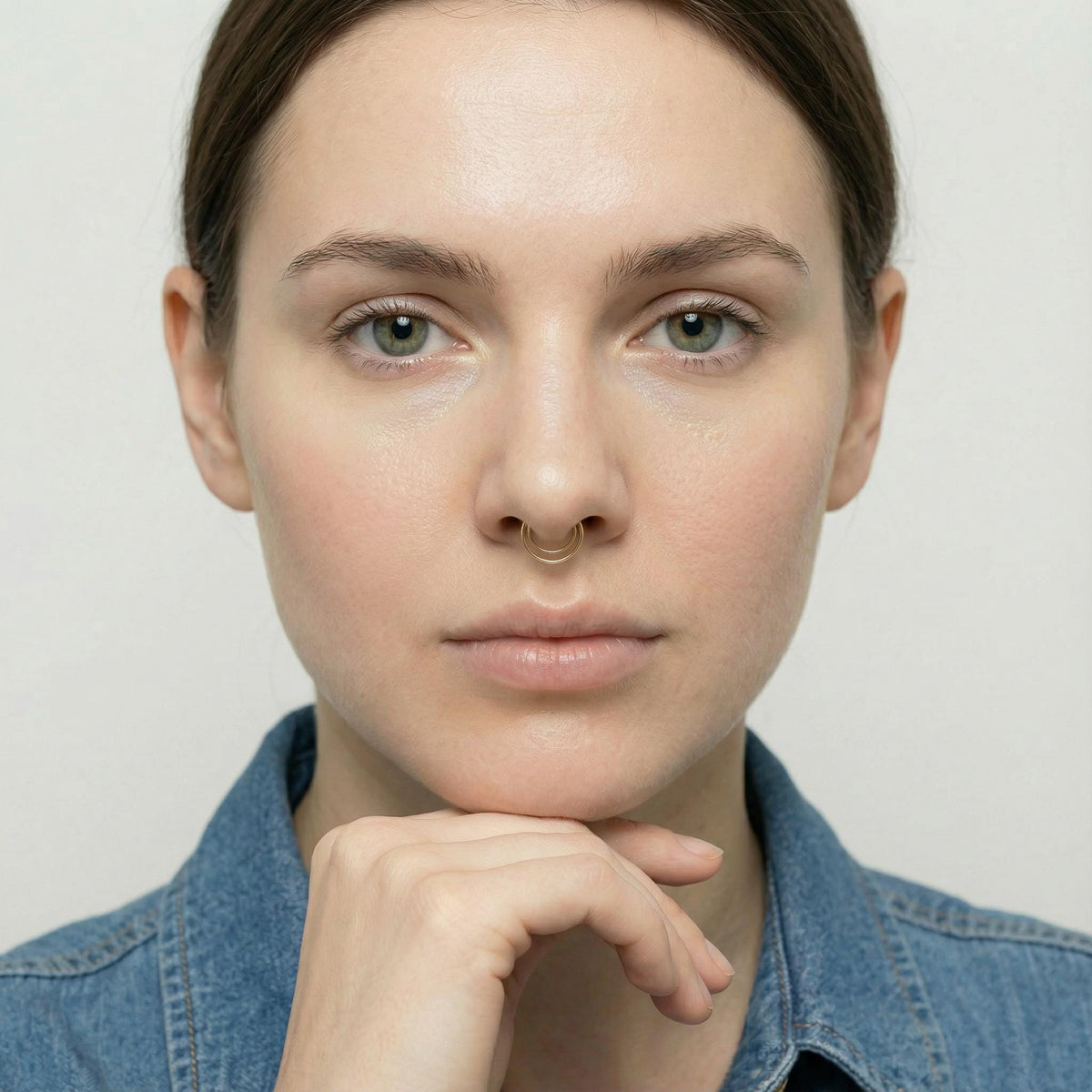 A close-up portrait of a woman with a thoughtful expression, wearing a denim jacket and a nose ring.