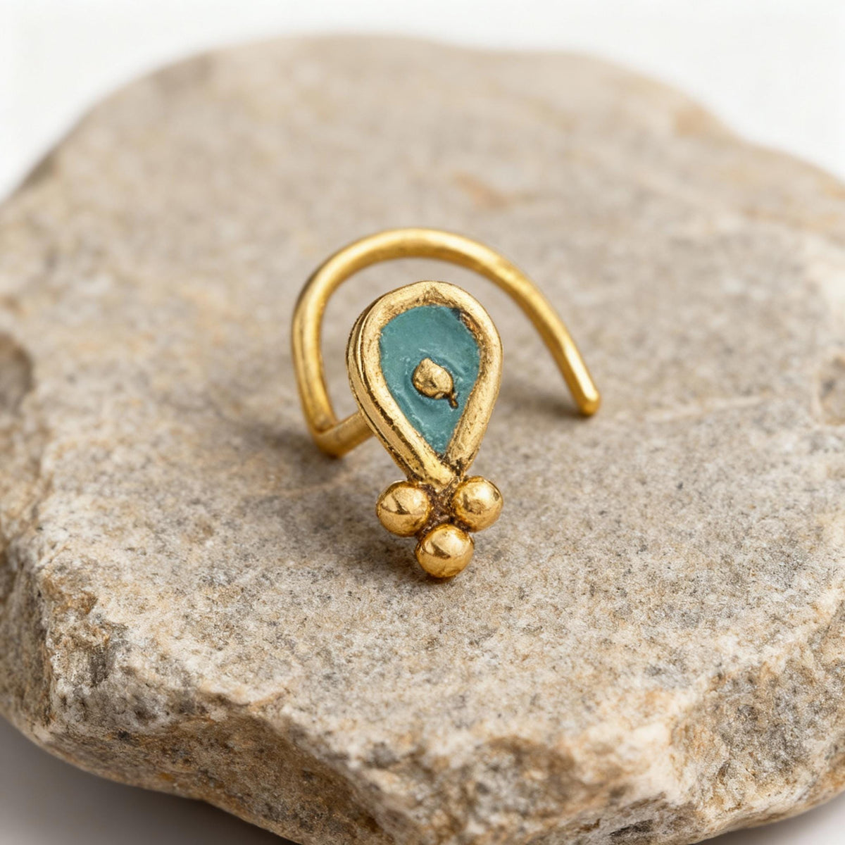 A gold-colored nose ring with a turquoise stone in the center, resting on a gray rock.