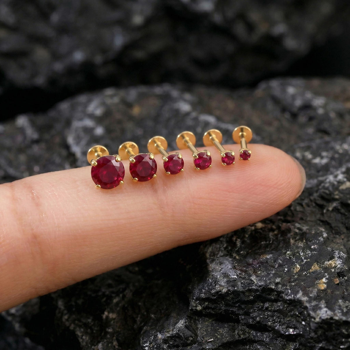 A close-up image of a person's finger wearing a row of gold-colored earrings with red gemstones.
