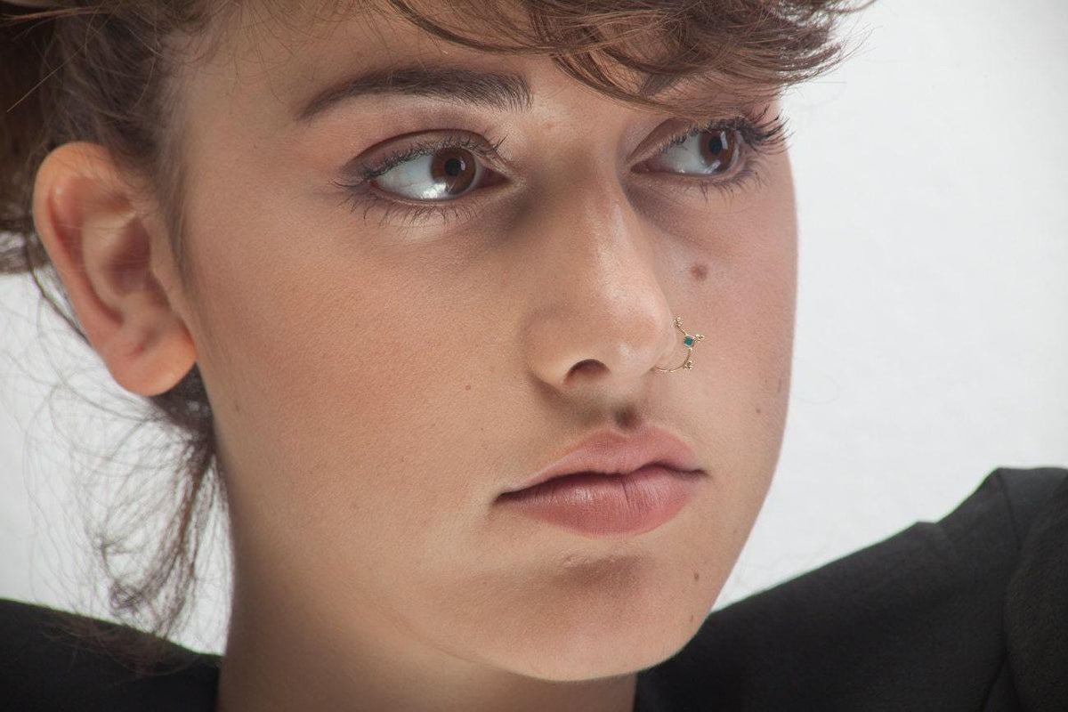 A close-up portrait of a young woman with a pensive expression, wearing a black top and a nose ring.