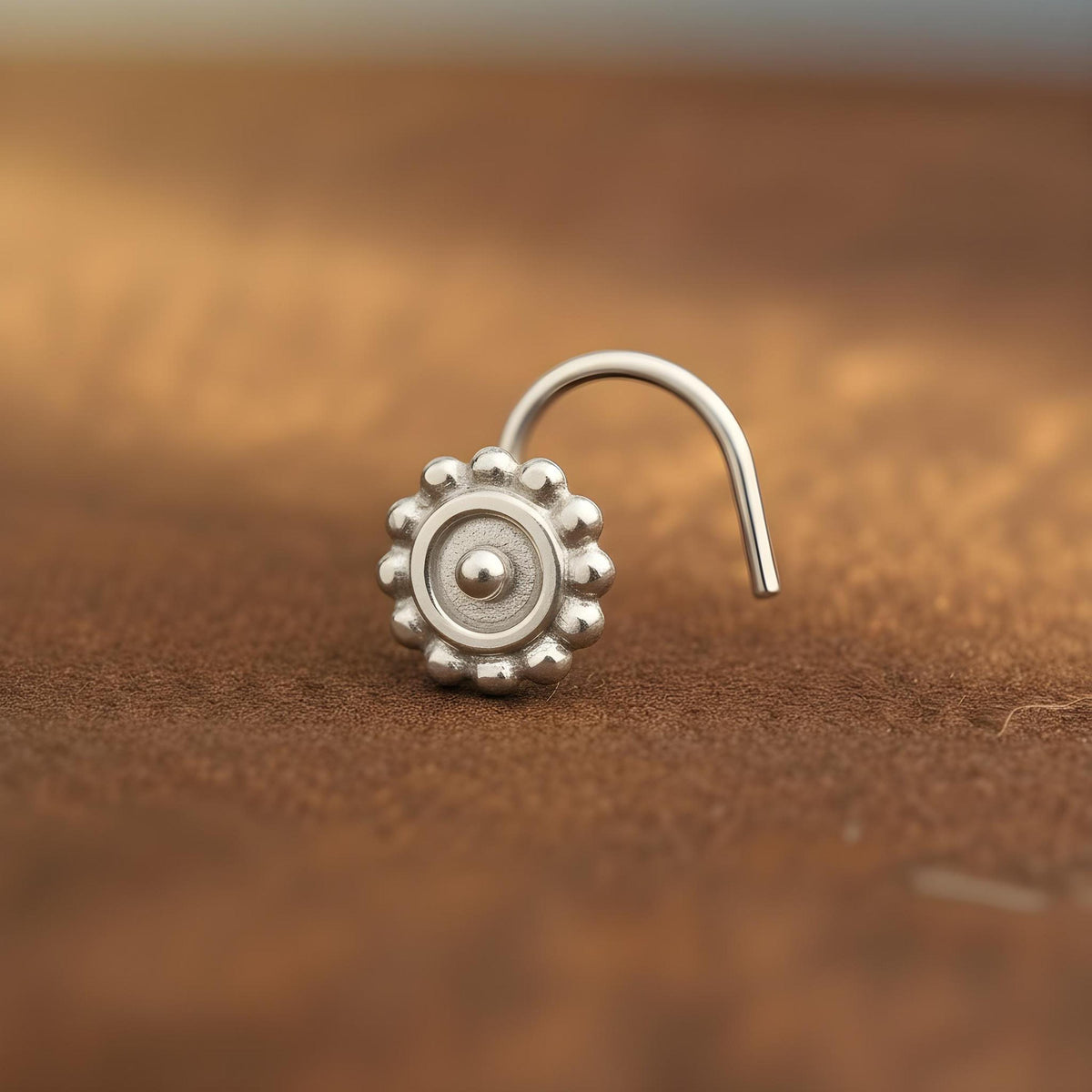 A silver nose stud with a flower-like design is shown against a blurred brown background.