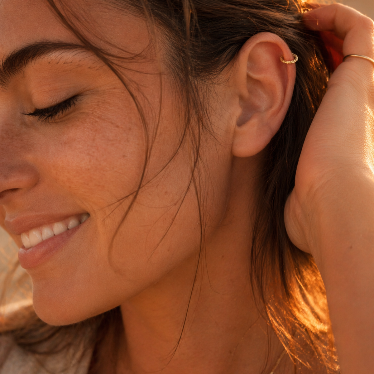 A close-up of a woman's face, with her eyes closed and her hair styled in loose waves. She is smiling and has a gold earring in her ear.