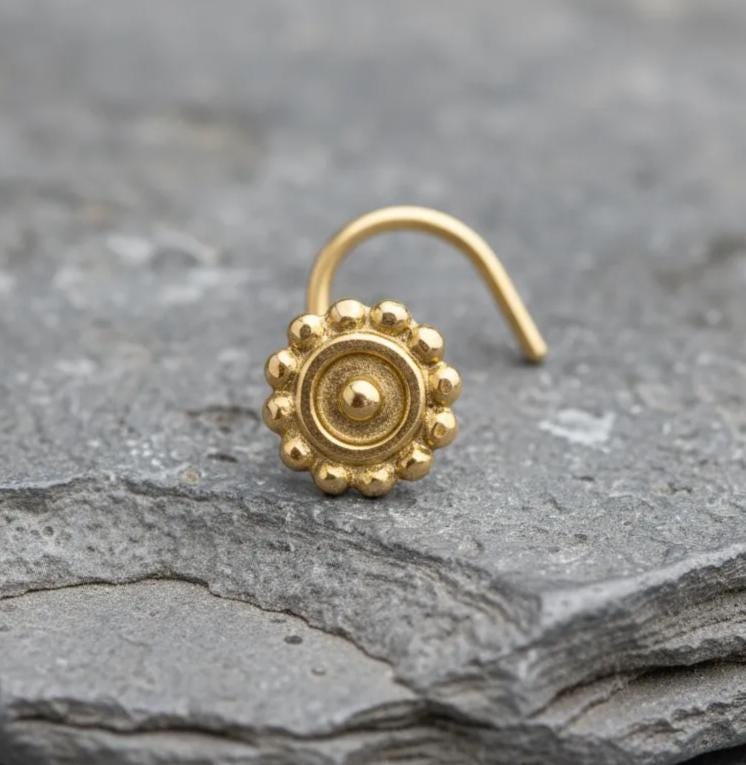 A gold-colored nose stud with a flower-like design is resting on a gray rock surface.