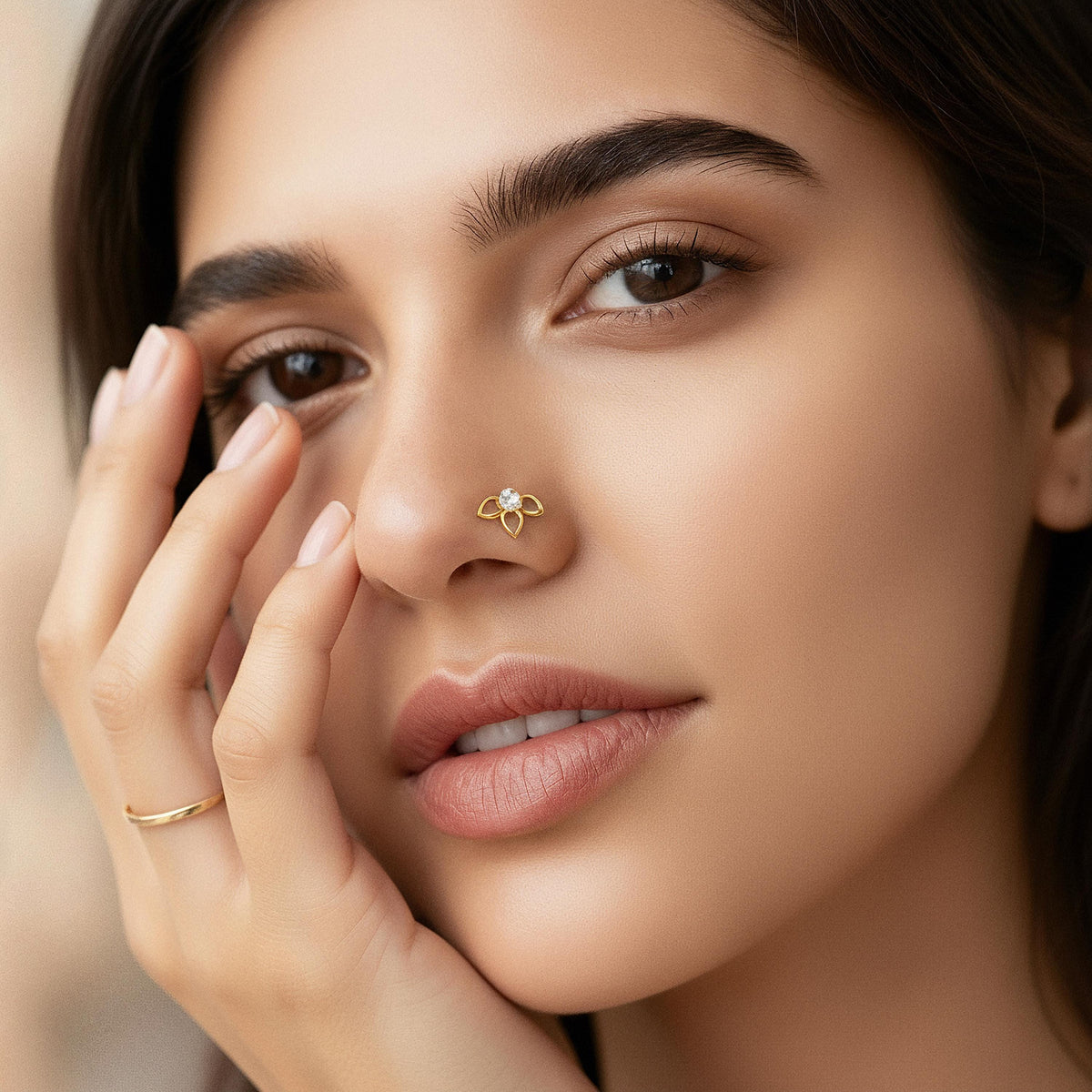 A close-up portrait of a woman with dark hair, wearing a gold nose ring and a ring on her finger.