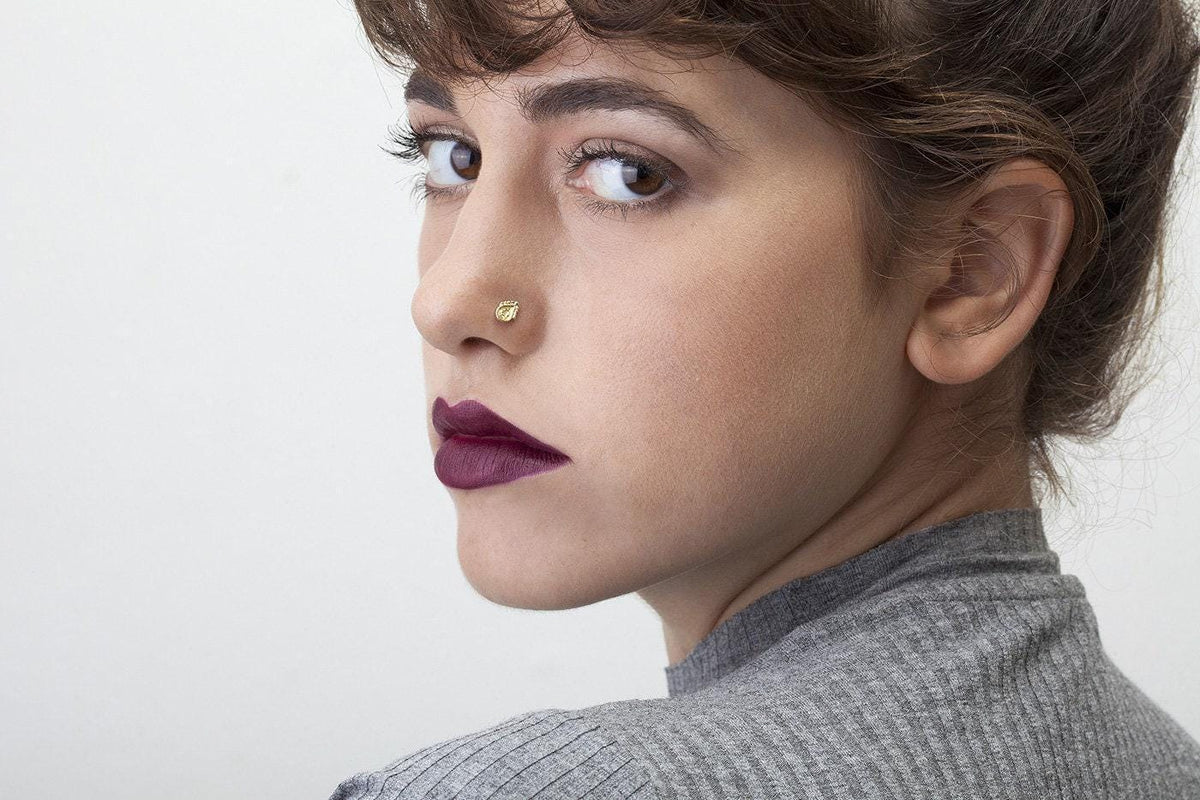 A close-up portrait of a woman with dark hair and a nose piercing, wearing a gray sweater.