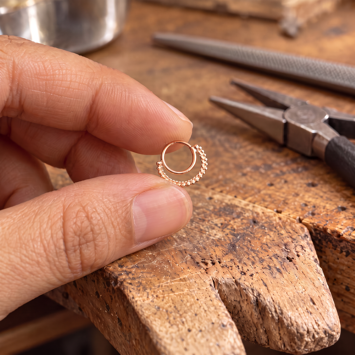 A hand holding a small gold-colored ring on a wooden surface, with various tools and materials visible in the background.
