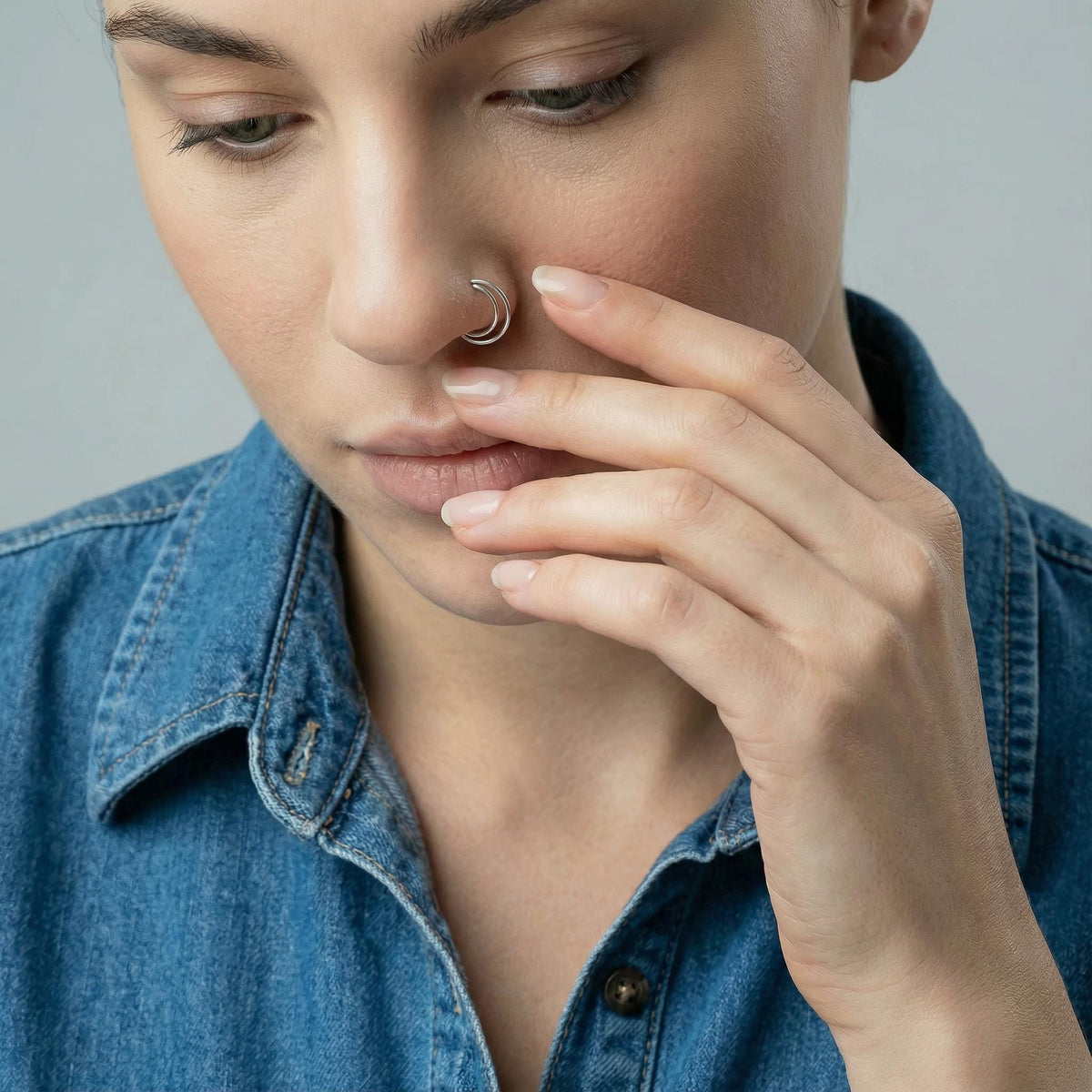 A close-up portrait of a woman wearing a denim shirt, with her hand near nose and mouth. She wears white gold double hoop ring.