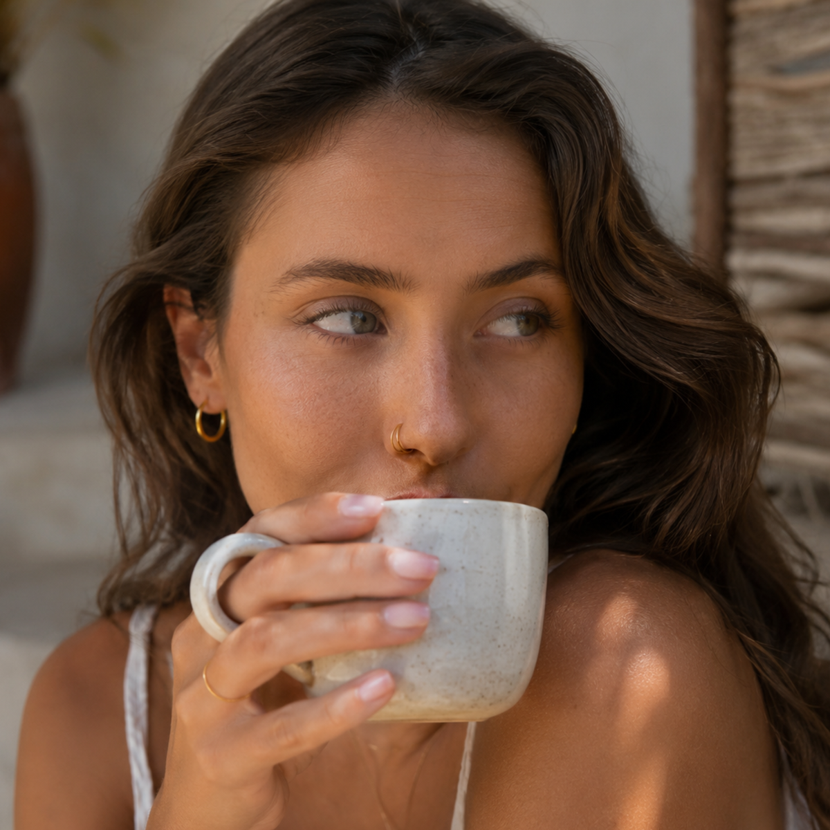 A woman with long brown hair is holding a white mug and sipping from it, looking pensively to the side.