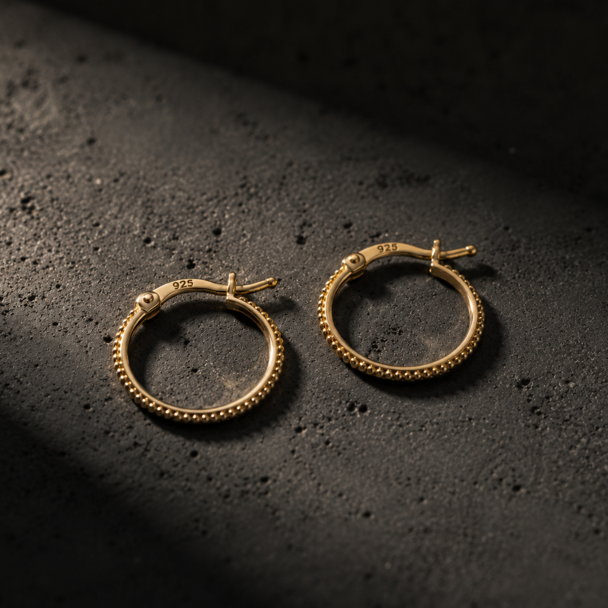 Two gold earrings with a textured design are displayed on a dark surface.